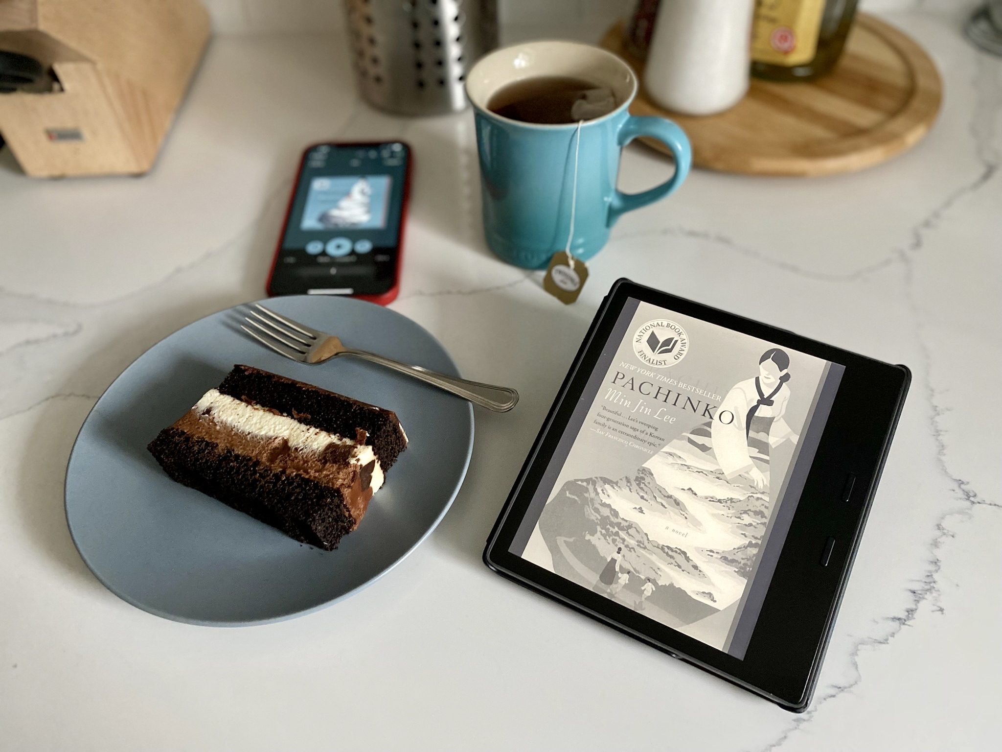 The cover of the book Pachinko both in a Kindle device and a cell phone, a cup of tea and a slice of chocolate cake over a kitchen marble countertop.