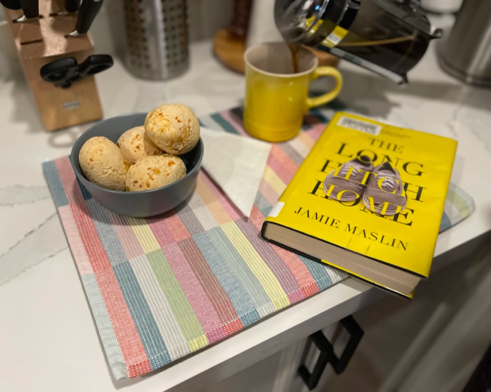 The Long Hitch Home by Jamie Maslin, over the kitchen counter by a bowl of Brazilian cheese bread and coffee being poured on a yellow cup. A book with a map.