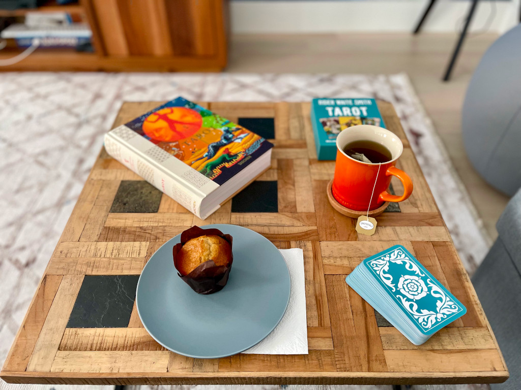 A coffee table laid with the book Tarot, The Library of Esoteric, an orange tea cup, a Tarot deck and a banana muffing in a desert plate over a napkin. A carpet an some furniture in the background.